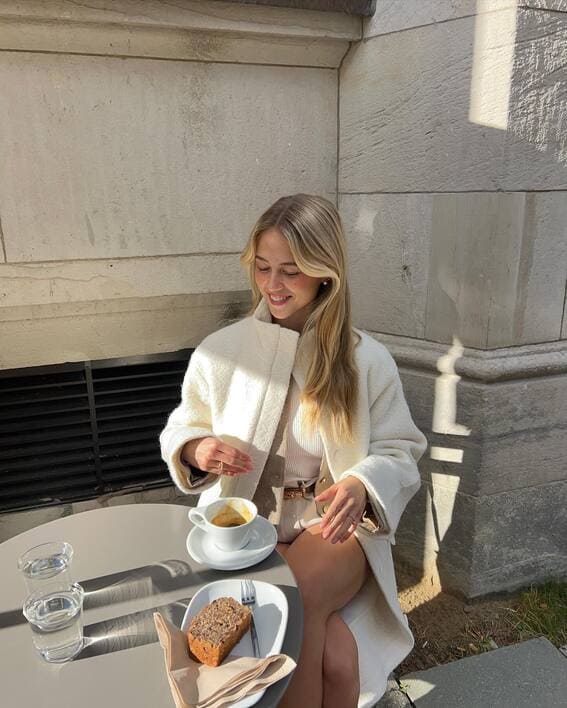 A woman is wearing an off-white coat with neutral-toned clothes while drinking coffee and banana bread at an outdoor café.