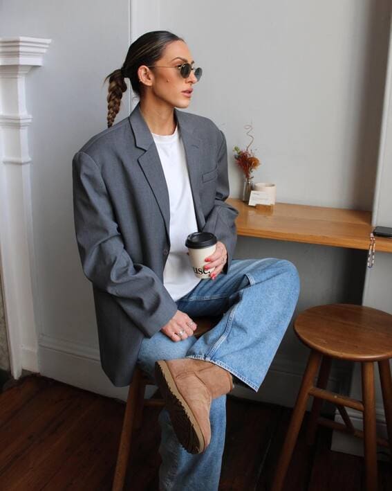 A woman wearing an oversized gray blazer, a white T-shirt, Light-wash jeans, and Ugg boots with a tan hue is sipping coffee at a café.