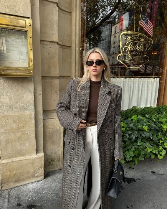 Woman in a herringbone overcoat with tailored white trousers, standing outside Ralph’s Coffee. 