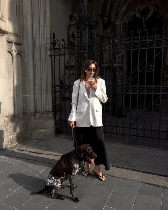 A woman in a white pinstripe blazer and black skirt is holding a drink while walking her dog. 