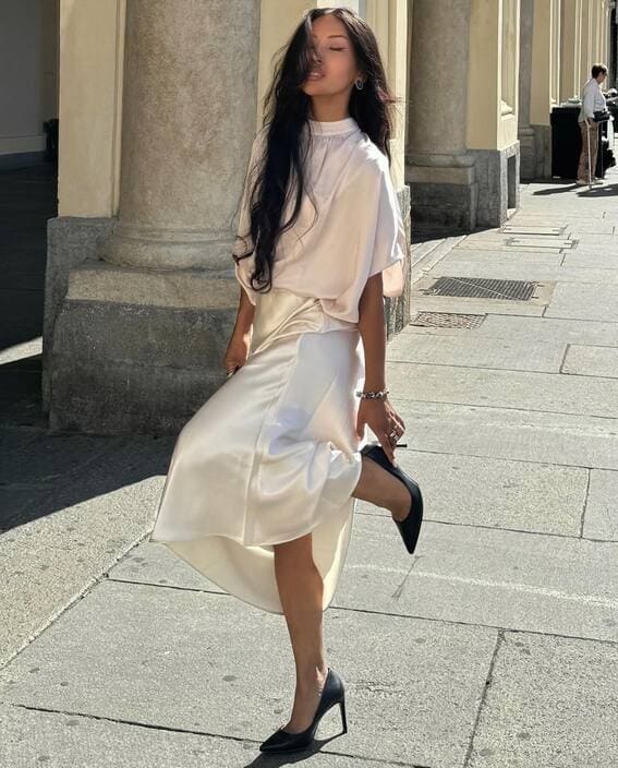 A woman in a cream silk blouse and satin skirt stands with one leg lifted in a playful pose. 