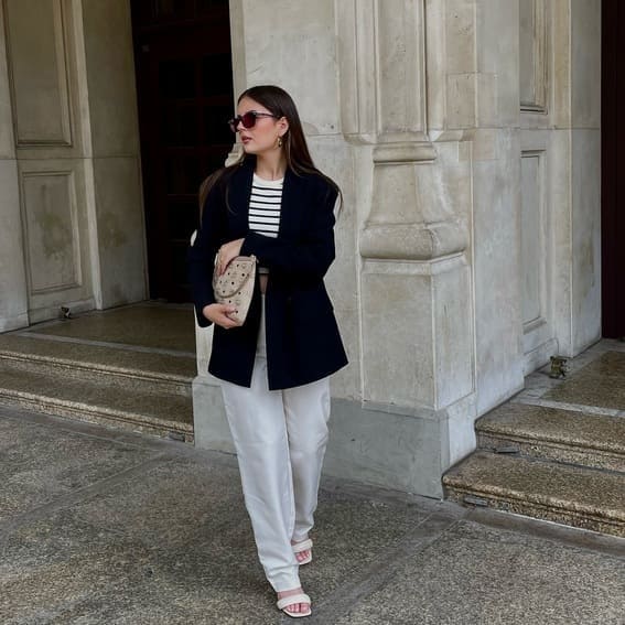 A woman in a striped top, navy blazer, and white trousers stands near a stone building. 