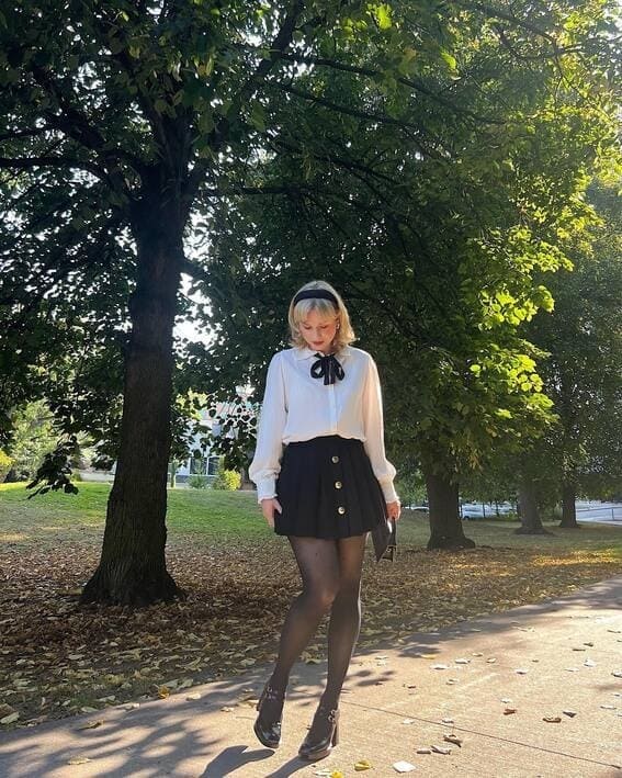 A woman in a white blouse with a black bow and pleated skirt stands in a sunlit park. 