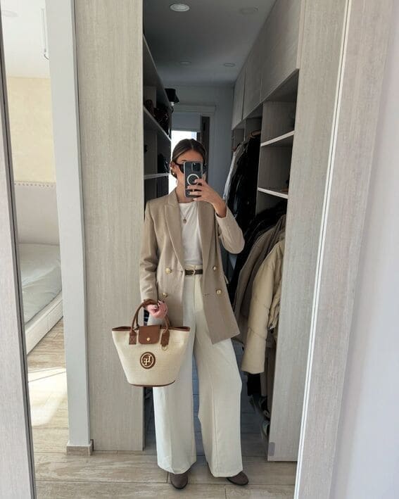 A woman in a beige double-breasted blazer and white wide-leg trousers is taking a mirror selfie in a chic walk-in closet. 