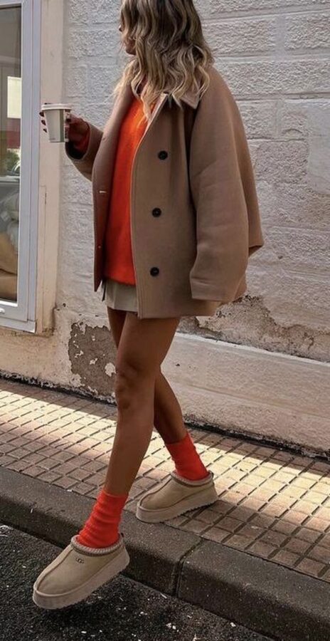 Woman wearing a camel oversized coat, bright orange sweater, mini skirt, orange socks, and beige mini UGG boots
