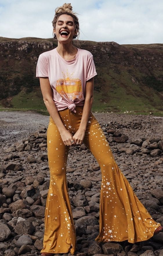 Woman wearing mustard star-print flared pants and a tied pink graphic tee, laughing outdoors on rocky terrain