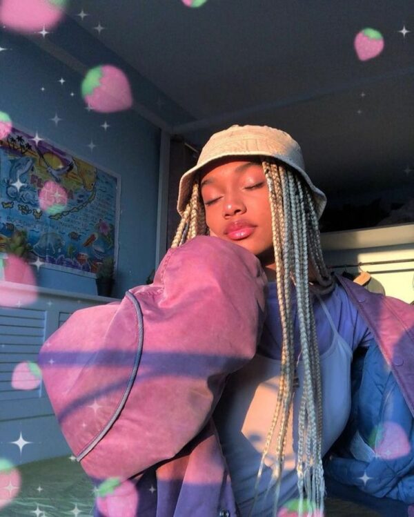 A woman with long, blonde box braids wears a neutral bucket hat and a lavender oversized jacket, surrounded by pink strawberry and sparkly heart overlays.