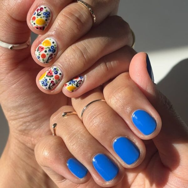 Short nails with a pop of vibrant blue paired with colorful fruit-themed designs on a creamy white base.