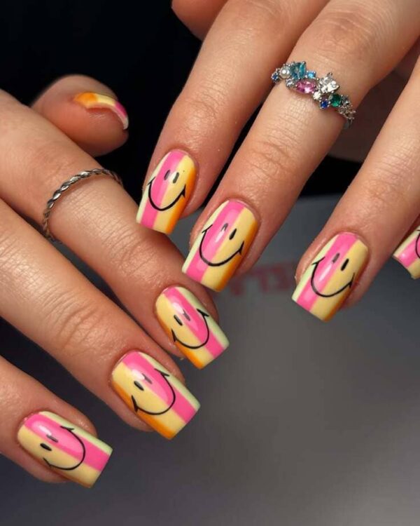 Playful yellow, pink, and orange striped nails featuring cute black smiley face designs on a glossy finish.
