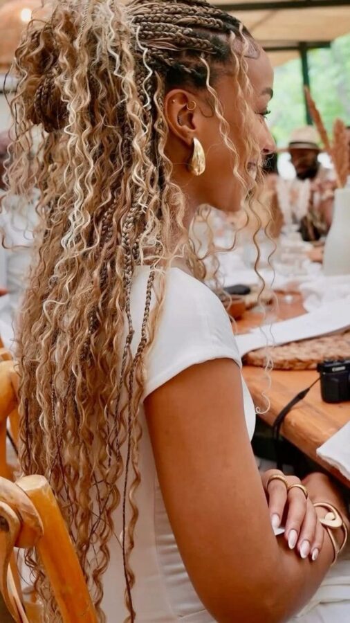 Woman with boho braids and long curly hair styled in a half-up look, wearing gold earrings and a white top