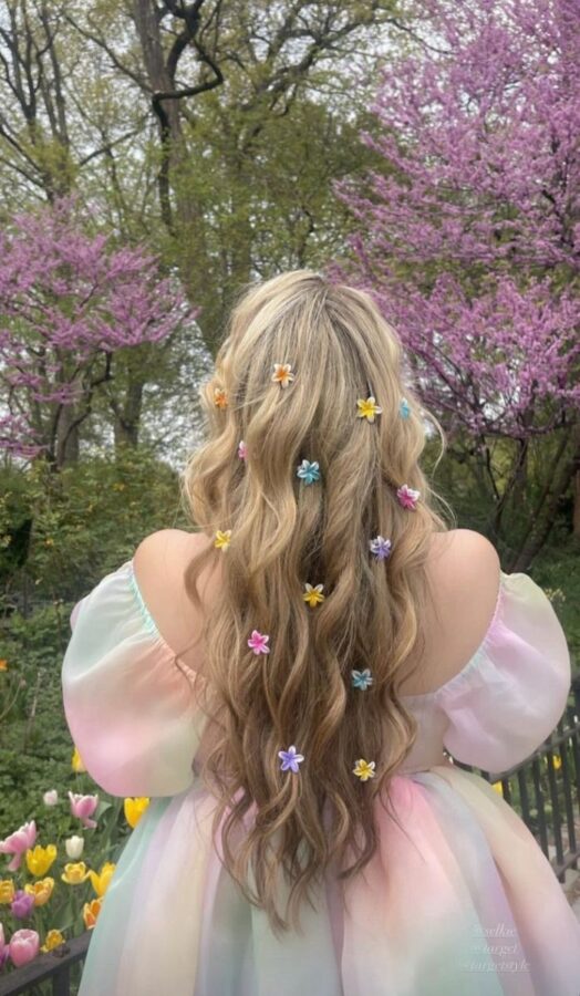 A long wavy hairstyle with loose curls, decorated with colorful pastel flower clips, worn with an off-shoulder pastel rainbow dress in a blooming spring garden.