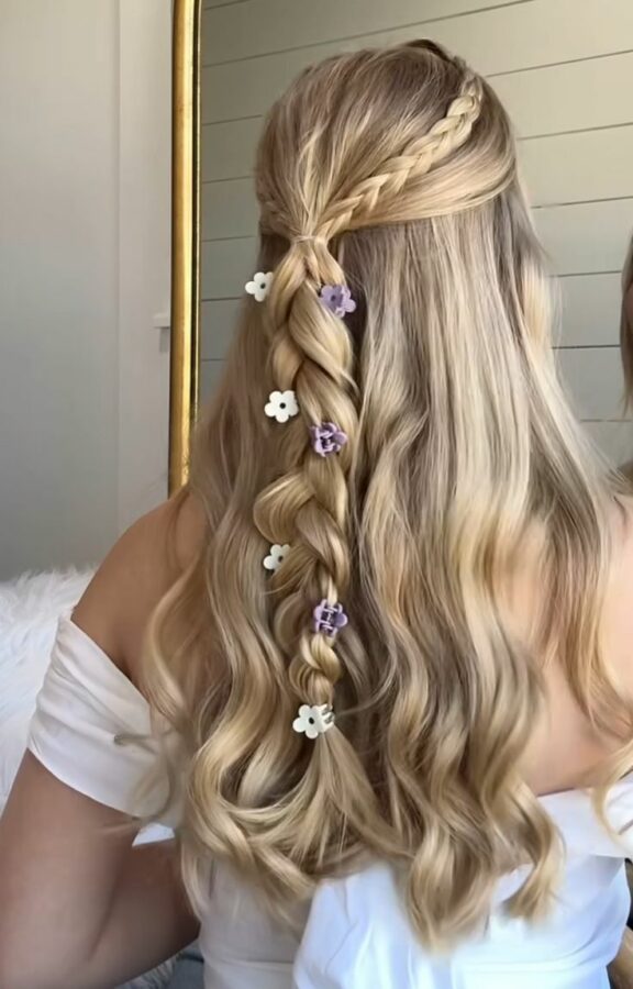 A long, wavy blonde hairstyle featuring a loose braid decorated with small white and purple flower clips, paired with a side braid and soft curls.