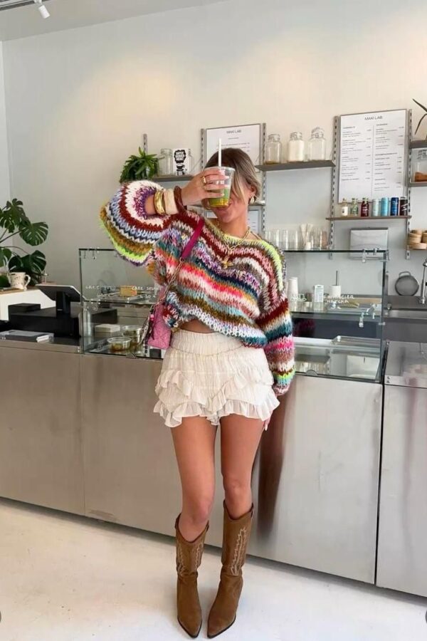 Woman wearing a rainbow knit sweater with ruffled cream shorts and brown boots in a café