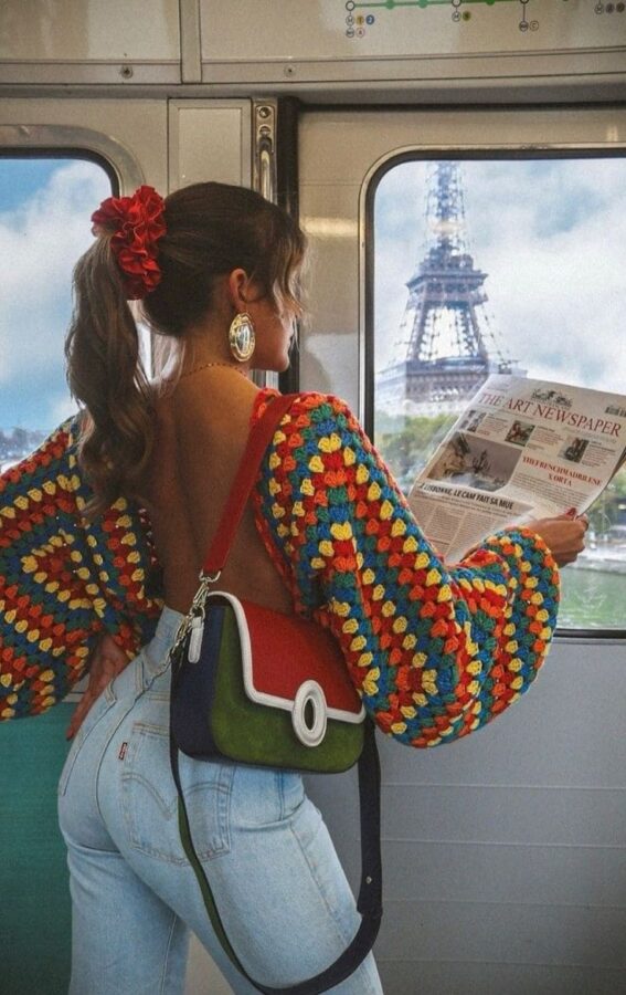 Woman wearing a vibrant open-back crochet sweater with light-wash jeans, red scrunchie, and color-block crossbody bag on a train with the Eiffel Tower in view
