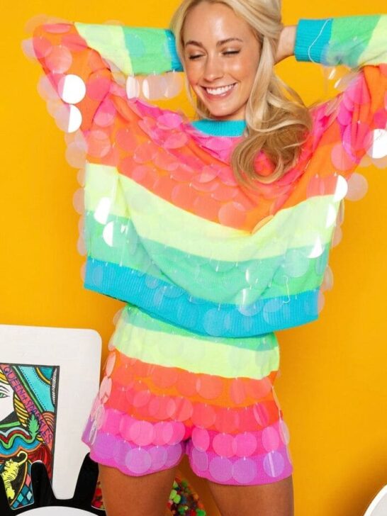 A woman wearing a neon rainbow striped sweater and matching shorts, covered in oversized clear sequins, smiling and posing against a bright yellow wall.