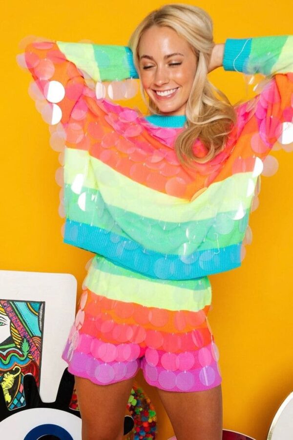 A woman wearing a neon rainbow striped sweater and matching shorts, covered in oversized clear sequins, smiling and posing against a bright yellow wall.
