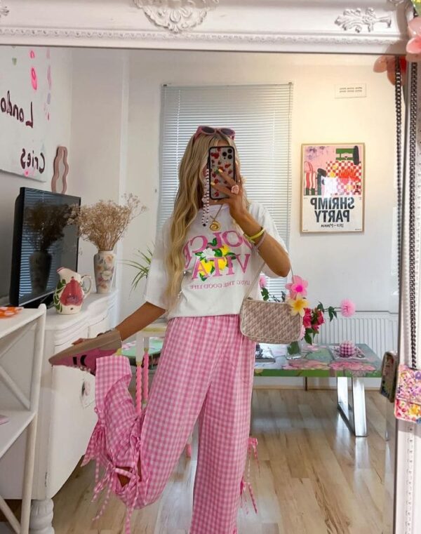 A woman wears a white graphic tee tucked into pink gingham wide-leg pants with ribbon details, paired with a beige crossbody bag and layered jewelry, posing in front of a mirror.