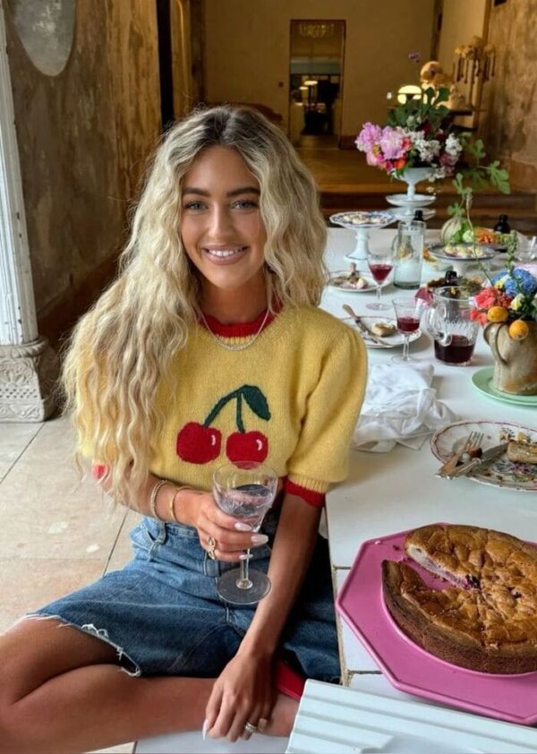 A woman with wavy blonde hair wears a yellow cherry-print knit sweater with a red collar, paired with a denim skirt and delicate jewelry, seated at a table full of brunch treats.