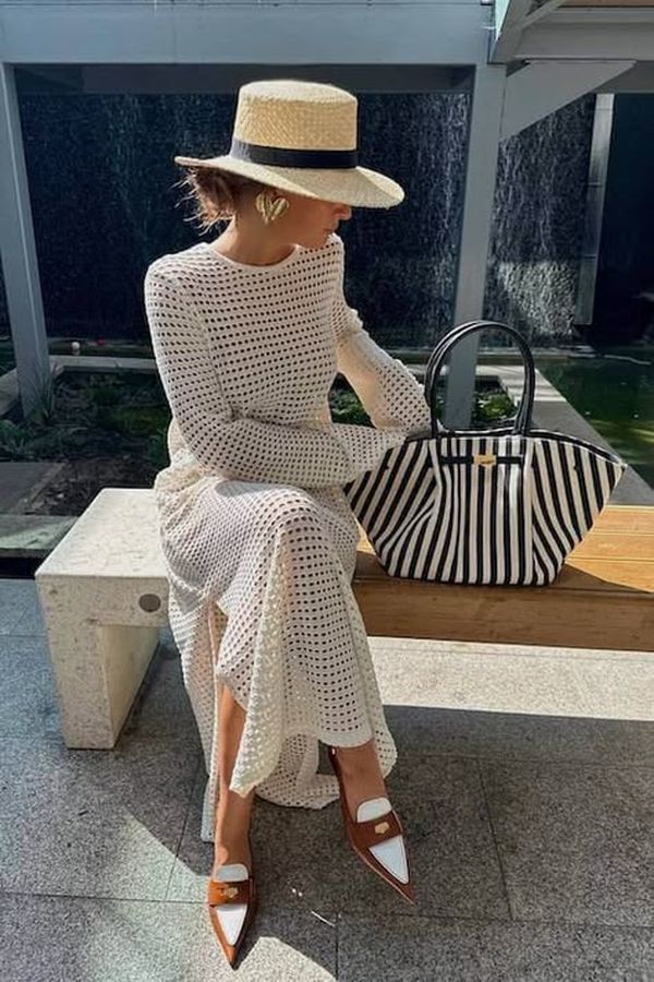 Woman wearing a white open-knit midi dress with a straw boater hat, gold statement earrings, brown pointed flats, and a structured black-and-white striped handbag